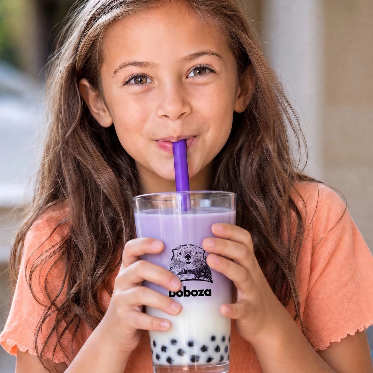 A young girl smiling as she sips a Boboza milk tea with boba pearls through a purple straw from a branded Boboza cup
