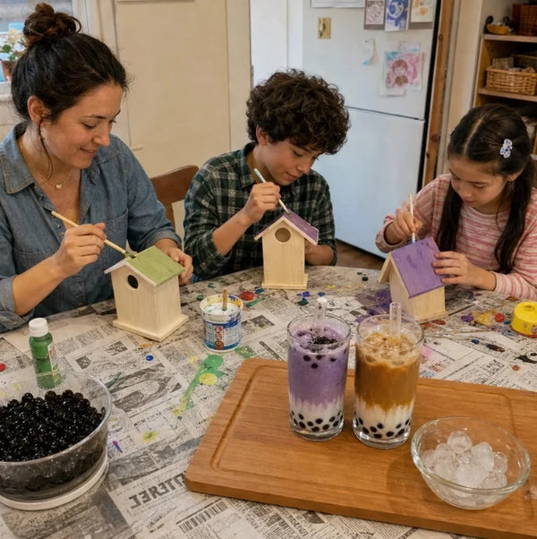 Mom and two kids painting wooden birdhouses at the kitchen table with two boba drinks and a bowl of boba pearls