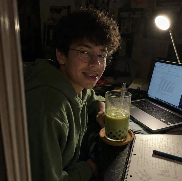 Teen studying late at his desk with a laptop, notebook and a matcha boba under a desk lamp