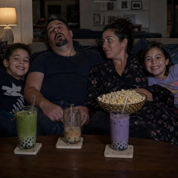 Family movie night on the couch with popcorn and three boba drinks on the coffee table, dad asleep while the kids laugh