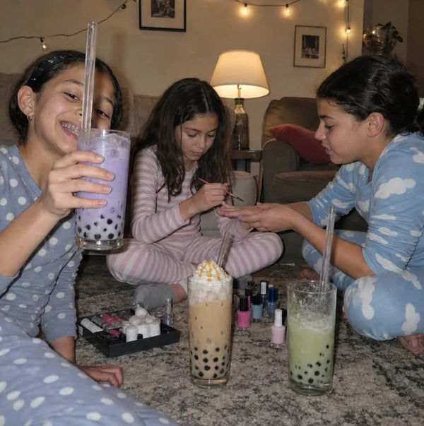 Three girls in pajamas painting their nails on the living room floor with three boba drinks beside them