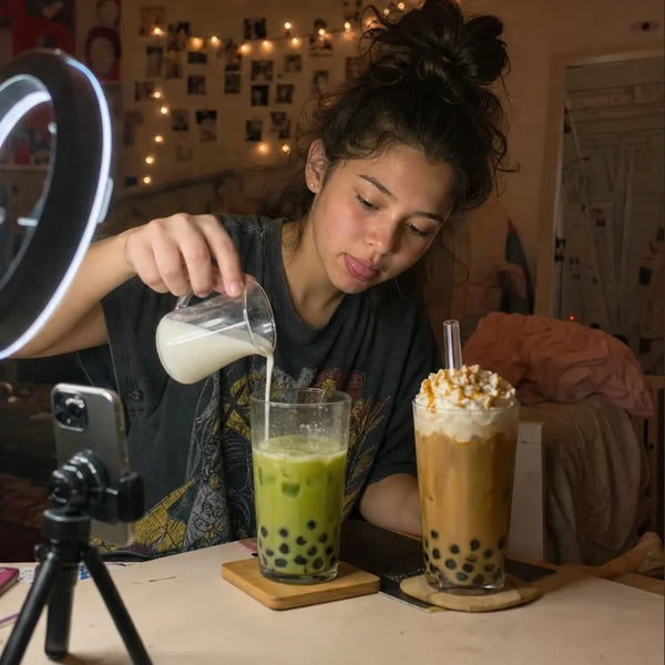 Teen filming herself pouring milk into a matcha boba with a ring light and phone tripod in her bedroom, a caramel whipped boba beside it