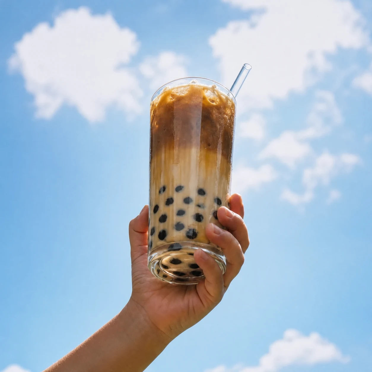 A hand holding up a tall glass of iced brown sugar boba milk tea with boba pearls at the bottom against a bright blue sky with white clouds